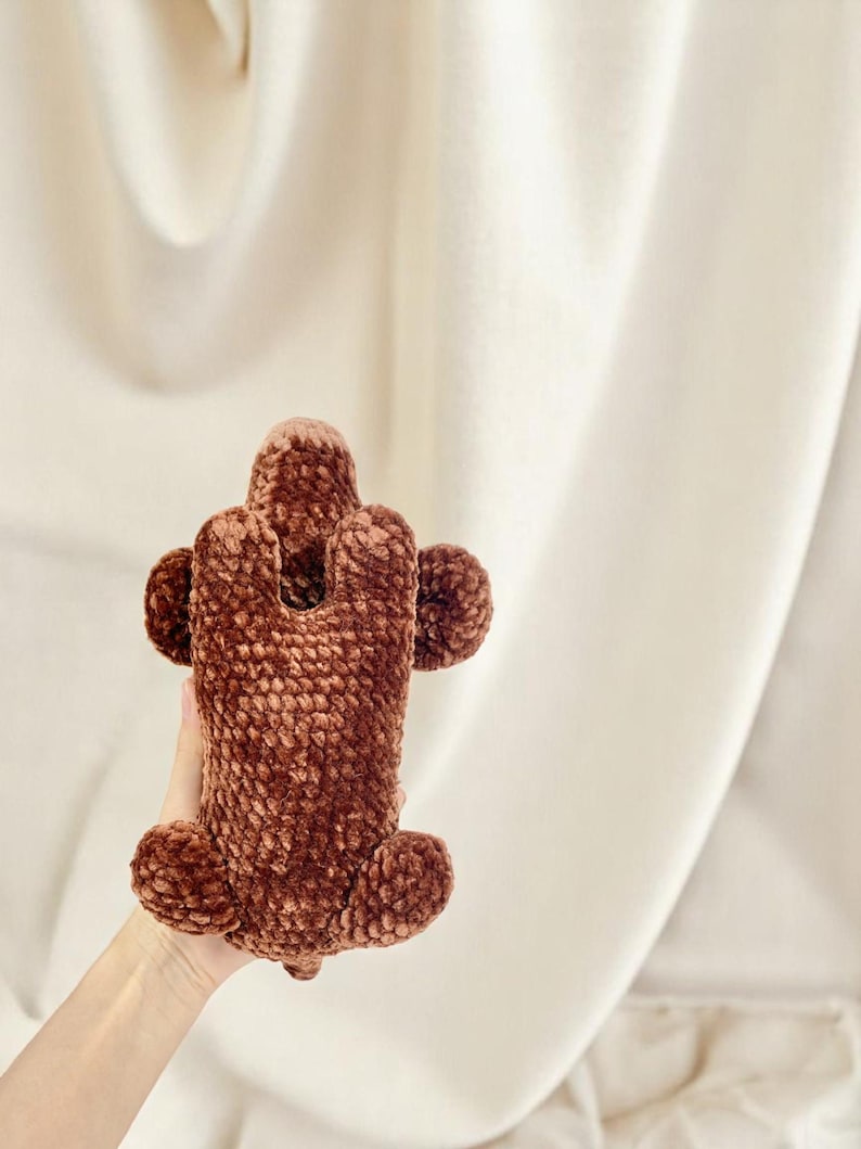 K&ouml;nnte beinhalten: Eine braune, pl&uuml;schige, geh&auml;kelte Handyh&uuml;lle. Die H&uuml;lle hat einen rechteckigen K&ouml;rper mit abgerundeten Kanten und kreisf&ouml;rmigen Akzenten. Die H&uuml;lle besteht aus einem weichen, strukturierten Material.