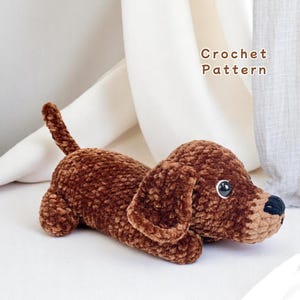 May include: A brown crocheted dachshund dog laying down. The plush toy has large floppy ears, a long body, and a small tail. The dog has black eyes and a black nose. The text "Crochet Pattern" is in the background.