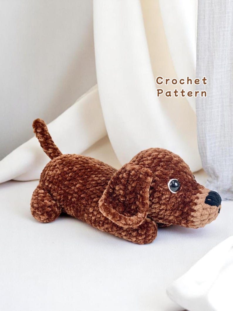 K&ouml;nnte beinhalten: Ein brauner geh&auml;kelter Dackelhund liegt. Das Pl&uuml;schtier hat gro&szlig;e Schlappohren, einen langen K&ouml;rper und einen kleinen Schwanz. Der Hund hat schwarze Augen und eine schwarze Nase. Der Text "Crochet Pattern" befindet sich im Hintergrund.