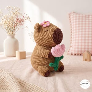 May include: A brown crocheted capybara plush toy featuring a dark brown snout, a small pink flower on its head, and holding a light pink crocheted flower with a green stem. The plush has closed eyes and dark brown feet, crafted with yarn.