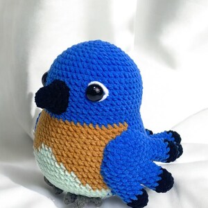Puede incluir: Un juguete de p&aacute;jaro de crochet azul, marr&oacute;n y verde con ojos negros y un pico negro. El p&aacute;jaro est&aacute; sentado sobre una superficie blanca.