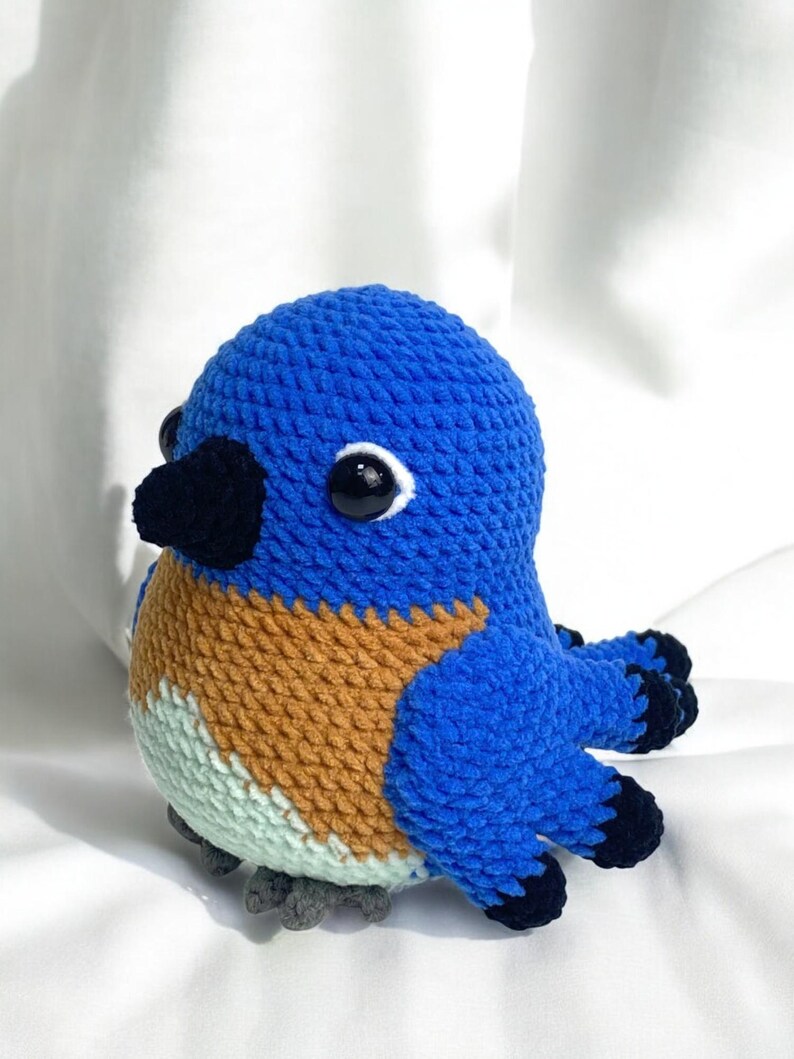 Puede incluir: Un juguete de p&aacute;jaro de crochet azul, marr&oacute;n y verde con ojos negros y un pico negro. El p&aacute;jaro est&aacute; sentado sobre una superficie blanca.