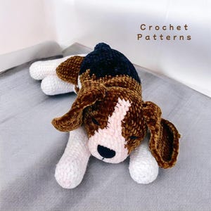 May include: A crocheted plush beagle dog toy in brown, white, and black. The dog is lying down with its eyes closed, as if sleeping.