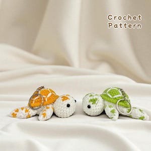 Peut inclure: Deux porte-clés en forme de tortue crochetées. L'une a une carapace orange et blanche, l'autre une carapace verte et blanche. Les deux ont des yeux noirs et un anneau de porte-clés argenté. Le texte "Crochet Pattern" est en haut à droite.