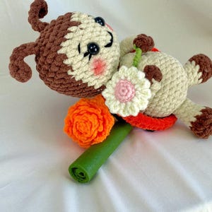 May include: A handmade crochet doll in a lying position, featuring a brown and cream-colored head and limbs. The doll holds a green stem with an orange and white flower. The doll has black button eyes and pink cheeks.