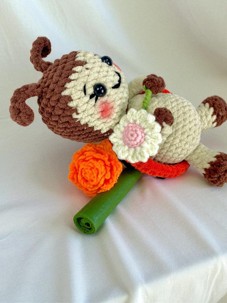 May include: A handmade crochet doll in a lying position, featuring a brown and cream-colored head and limbs. The doll holds a green stem with an orange and white flower. The doll has black button eyes and pink cheeks.