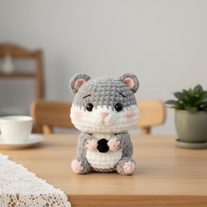 May include: A handmade crochet hamster toy in gray, white, and pink. The plush animal has black eyes, pink ears, and a small black object held in its paws. The toy is sitting on a wooden surface.