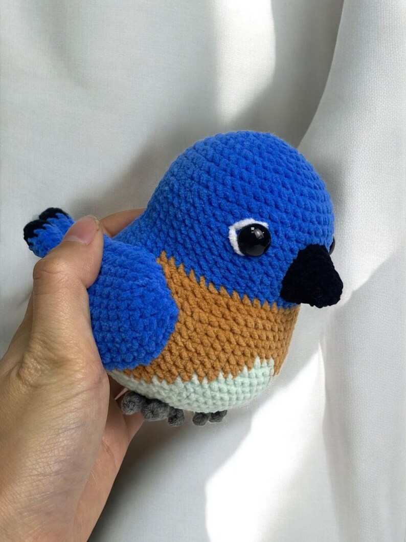 Puede incluir: Un juguete de peluche de p&aacute;jaro azul tejido a crochet con detalles marrones y blancos. El p&aacute;jaro tiene ojos negros y un pico negro.