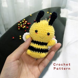 Può includere: Un giocattolo amigurumi a forma di ape realizzato all'uncinetto con strisce nere e gialle, ali bianche e antenne nere. L'ape ha un viso sorridente con occhi neri e un blush rosa. Il giocattolo è tenuto in una mano. Schema all'uncinetto.