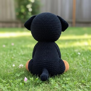 Puede incluir: Un peluche de perro negro hecho a mano, visto desde atr&aacute;s, sentado sobre hierba verde. El perro tiene patas marrones y una peque&ntilde;a cola. Las orejas son ca&iacute;das. El fondo est&aacute; borroso, lo que sugiere un entorno al aire libre.