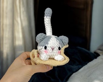 May include: A crocheted gray and white cat toy with pink cheeks sitting on a crocheted yellow banana. The cat toy has a long gray and white tail.