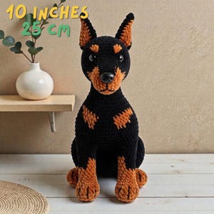 Op de afbeelding: Een handgemaakte zwart-bruine gehaakte Dobermann Pinscher hond, ongeveer 25 cm hoog. De hond heeft een gedetailleerd ontwerp met oranje accenten op de oren, borst en poten. De hond zit rechtop.
