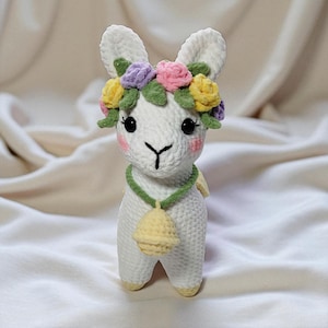 Puede incluir: Una llama blanca hecha a mano con ganchillo que lleva una corona floral con flores rosas, moradas y amarillas. La llama tiene ojos negros, mejillas rosas y un collar verde con una campana amarilla. La llama está sobre una superficie de tela blanca.