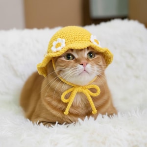 May include: A ginger cat wearing a yellow crochet hat decorated with white daisy flowers. The hat has a wide brim and a yellow chin strap tied in a bow. The cat is lying on a fluffy white surface. The hat is handmade.