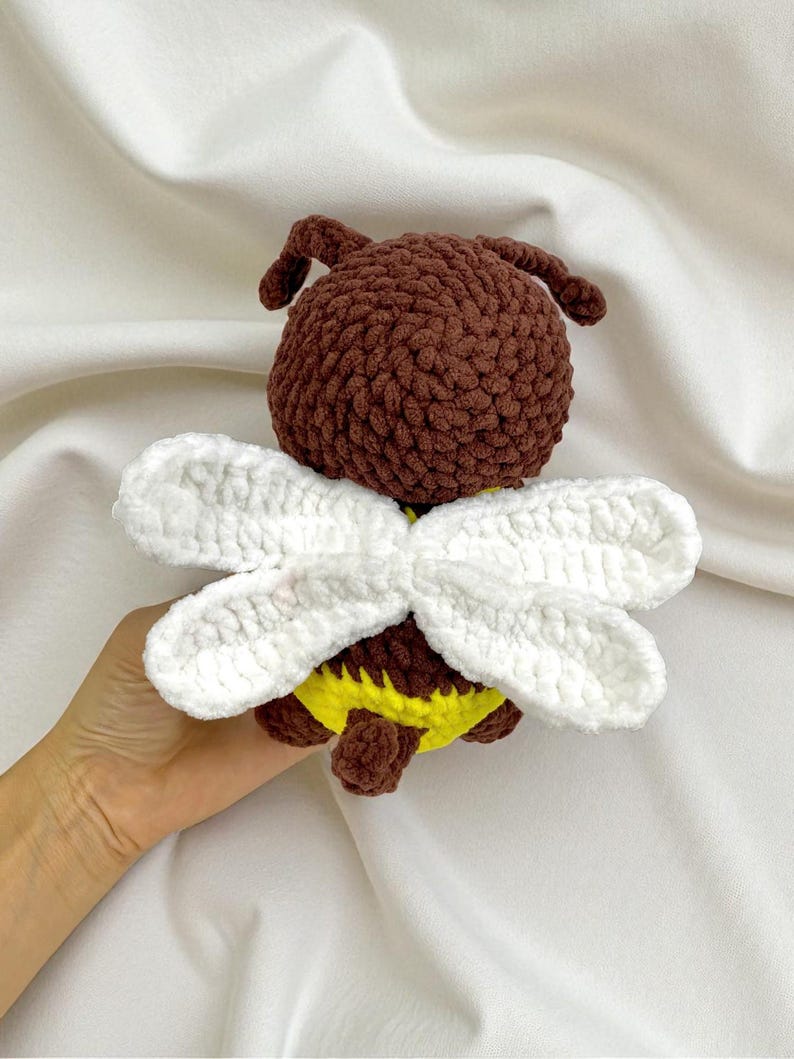 May include: A handmade crochet bee toy. The bee has a brown body, white wings, and a yellow and brown abdomen. The toy is made of soft yarn and is designed to be a cute and cuddly item.