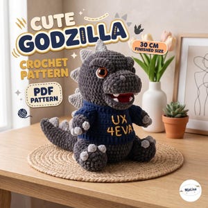 May include: A dark gray crocheted Godzilla plush toy wearing a navy blue sweater with the yellow text UX 4EVA. The plush features light gray dorsal fins, large brown eyes, and a wide mouth with white teeth. The text indicates a 30 cm finished size.