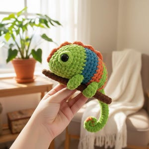 Puede incluir: Un camaleón de ganchillo hecho a mano en tonos verdes, azules y naranjas, posado en una ramita marrón. El peluche tiene grandes ojos negros y una cola enroscada. El camaleón es una creación caprichosa y colorida.