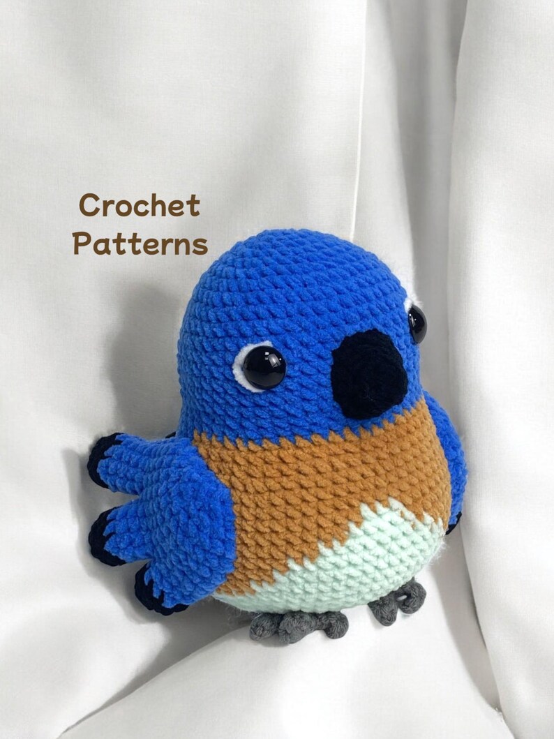 Puede incluir: Un juguete de peluche de p&aacute;jaro azul tejido a crochet con detalles marrones, blancos y azul claro. El p&aacute;jaro tiene ojos negros y un pico negro.