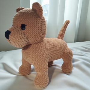 May include: A handmade, tan crochet dog with a black nose and eyes. The plush toy is in a standing pose with its tail up. The dog has a soft, textured appearance, and is likely a children's toy or decorative item. The dog is made of a soft yarn.