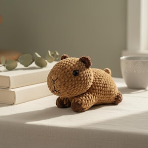 Könnte beinhalten: Handgehäkeltes Capybara in Brauntönen. Das Stofftier hat einen runden Körper, kleine dunkle Augen und dunkelbraune Akzente an Ohren und Füßen. Es liegt auf einer weißen Oberfläche.