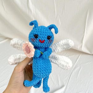 May include: A handmade blue crochet fairy doll with white wings and a pink flower. The doll has a smiling red mouth and black eyes. The doll is holding a pink flower and has two small antennae.