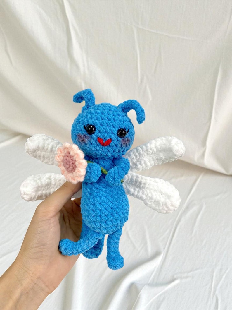 May include: A handmade blue crochet fairy doll with white wings and a pink flower. The doll has a smiling red mouth and black eyes. The doll is holding a pink flower and has two small antennae.