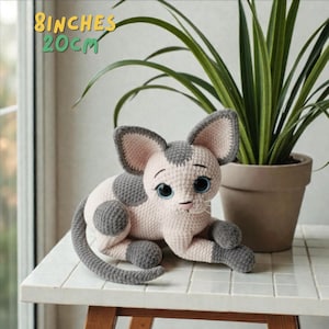 Peut inclure: Poupée chat au crochet faite à la main, rose et grise, avec de grands yeux bleus. Le chat est allongé, la queue enroulée. Le texte "8 inches 20cm" est dans le coin supérieur gauche. La poupée mesure 20 cm.