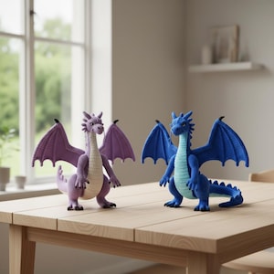 May include: Two handmade crochet dragons in shades of purple and blue sit on a wooden table. The dragons have detailed wings, bodies, and tails. The purple dragon has lavender wings, while the blue dragon has darker blue wings. The dragons are a whimsical decorative item.