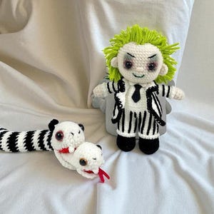 May include: Crocheted figures of a character with green hair, wearing a black and white striped suit, and a snake with black and white stripes. The snake has a white head with red accents.