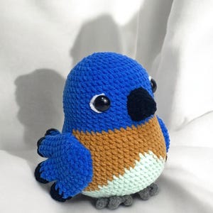 Puede incluir: Un juguete de peluche de p&aacute;jaro tejido a crochet azul y marr&oacute;n con ojos negros y un pico negro. El p&aacute;jaro est&aacute; sentado sobre una superficie blanca.
