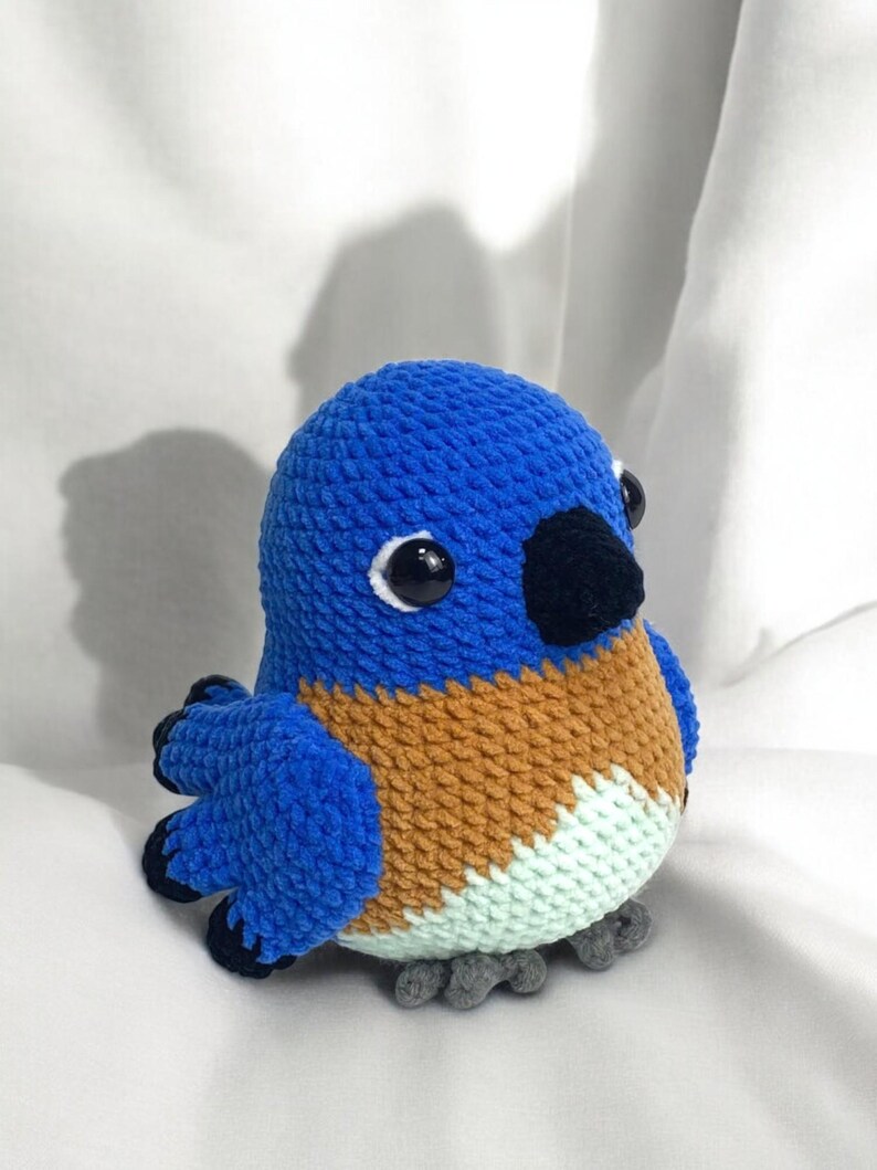 Puede incluir: Un juguete de peluche de p&aacute;jaro tejido a crochet azul y marr&oacute;n con ojos negros y un pico negro. El p&aacute;jaro est&aacute; sentado sobre una superficie blanca.