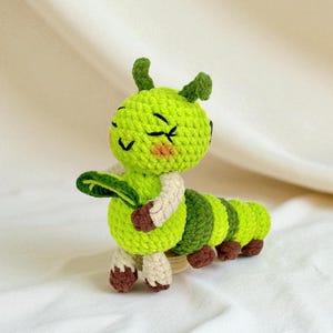 May include: A handmade crochet caterpillar toy in shades of green, brown, and cream. The caterpillar has a smiling face with closed eyes, a leaf, and segmented body. The toy is made of soft yarn and is likely a children's toy or decorative item.
