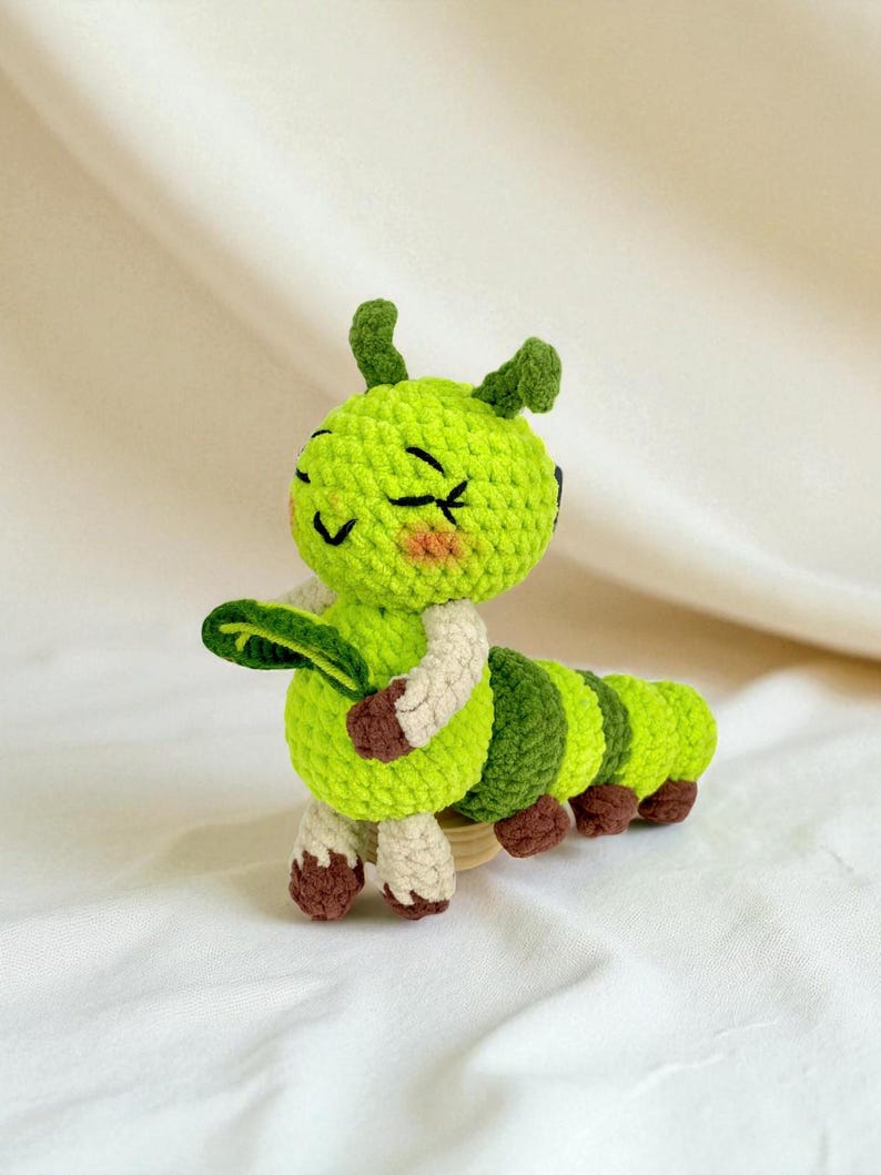 May include: A handmade crochet caterpillar toy in shades of green, brown, and cream. The caterpillar has a smiling face with closed eyes, a leaf, and segmented body. The toy is made of soft yarn and is likely a children's toy or decorative item.