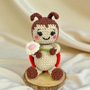 May include: A handmade crochet ladybug doll with a smiling face, holding a white and pink flower. The doll has a red back, brown antennae, and is sitting on a wooden slice. The doll is made of beige, brown, and red yarn.