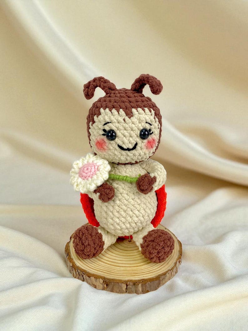 May include: A handmade crochet ladybug doll with a smiling face, holding a white and pink flower. The doll has a red back, brown antennae, and is sitting on a wooden slice. The doll is made of beige, brown, and red yarn.