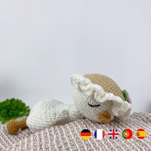 Puede incluir: Muñeca de ganchillo hecha a mano con forma de pájaro durmiente. El pájaro es principalmente blanco con cabeza y pico color beige. Lleva un sombrero blanco con borde con volantes y una pequeña flor. La muñeca está sobre una superficie beige texturizada.