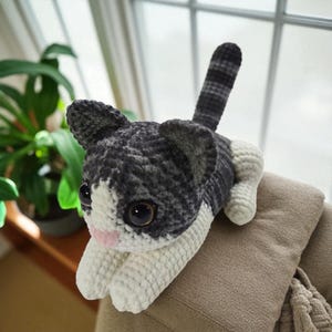 Può includere: Un peluche a forma di gatto fatto a mano all'uncinetto, grigio, bianco e nero. Il gatto ha grandi occhi scuri, un naso rosa ed è sdraiato su una superficie beige. La coda è a strisce e le orecchie sono dritte.