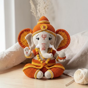 Peut inclure: Figurine de Ganesh crochetée, principalement orange, crème et jaune. La divinité à tête d'éléphant est assise, portant une couronne et des bijoux. Le fond comprend une surface en bois et un tissu de couleur neutre.