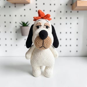 Puede incluir: Un juguete de peluche tejido a ganchillo con forma de perro, cuerpo blanco, orejas largas negras y hocico color canela. El perro tiene una nariz negra, ojos oscuros con párpados bordados y un pequeño accesorio naranja en la parte superior.