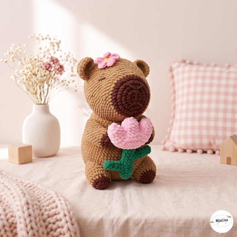 May include: A brown crocheted capybara plush toy featuring a dark brown snout, a small pink flower on its head, and holding a light pink crocheted flower with a green stem. The toy has closed eyes and a small logo tag reading MjaLisa.