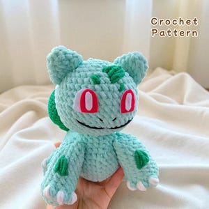 May include: A crocheted plush toy in the shape of a Bulbasaur, featuring a light teal body, red eyes, and green accents. The text "Crochet Pattern" is visible in the upper right corner. The toy is held in a hand.