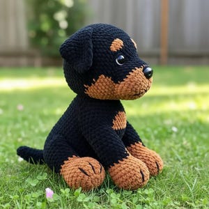 Puede incluir: Un juguete de perro hecho a mano con ganchillo, negro con marcas marrones, sentado en la hierba. El perro de juguete tiene una nariz negra y ojos grandes. Las marcas marrones est&aacute;n en el hocico, las patas y encima de los ojos.