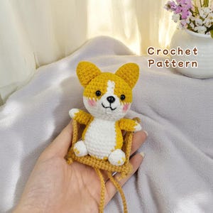 May include: A crocheted corgi dog plush toy sitting on a swing. The corgi is yellow and white with a brown nose and black eyes. The swing is made of brown yarn.