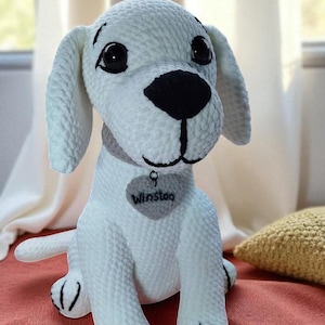 Puede incluir: Un perro de ganchillo blanco hecho a mano con detalles negros en la nariz, los ojos y las patas. El perro tiene orejas caídas y un collar gris con una etiqueta en forma de corazón que dice "Winston". El peluche está sentado sobre una superficie roja.