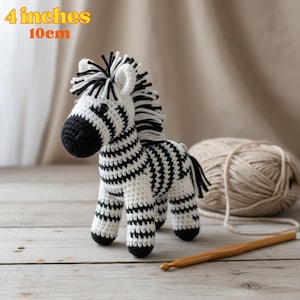 Może przedstawiać: Ręcznie robiona szydełkowa zebra, około 10 cm wysokości, z czarno-białymi paskami. Zebra ma czarną grzywę, ogon i pysk. Kłębek wełny i szydełko sugerują projekt rękodzielniczy.