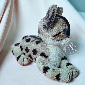 Peut inclure: Un chat en crochet fait à la main, en position couchée. Le chat est beige avec des taches marron foncé et des rayures noires. Il a des yeux bleus et des moustaches roses. Fabriqué en fil doux.