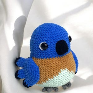 Puede incluir: Un juguete de peluche de p&aacute;jaro de crochet azul y marr&oacute;n con ojos negros y un pico negro. El p&aacute;jaro tiene un cuerpo azul claro, alas marrones y una barriga verde claro.