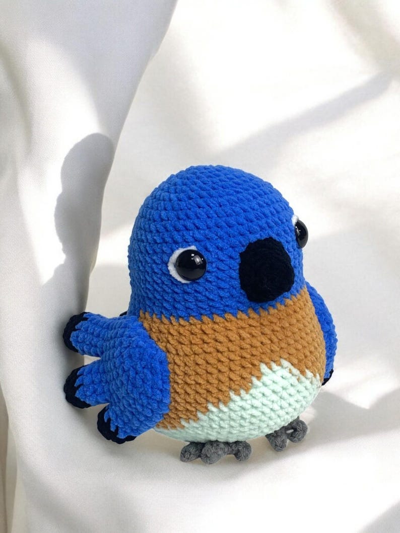 Puede incluir: Un juguete de peluche de p&aacute;jaro de crochet azul y marr&oacute;n con ojos negros y un pico negro. El p&aacute;jaro tiene un cuerpo azul claro, alas marrones y una barriga verde claro.
