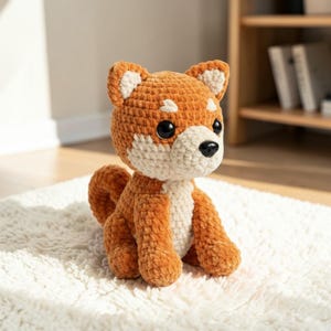 Op de afbeelding: Een handgemaakte gehaakte Shiba Inu hondenknuffel. Het pluche speelgoed is gemaakt van oranje en wit garen, met zwarte ogen en neus. De hond zit op een wit, pluizig tapijt. De oren en borst zijn wit, en de rest van het lichaam is oranje.