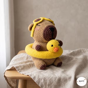 May include: A crocheted capybara plush toy featuring a tan body, dark brown snout, and pink cheeks. It wears a yellow aviator-style cap and is positioned inside a bright yellow crocheted swim ring shaped like a duck with a small orange beak.